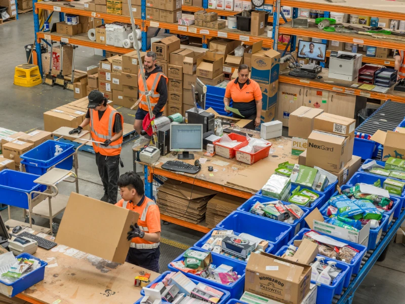 Busy warehouse packing area with staff in orange vests assembling orders among blue bins, boxes, and computer stations