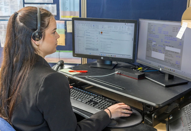 A customer service representative wearing a headset works at a desk with multiple computer monitors displaying emails and order information.