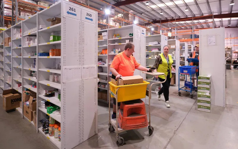 Warehouse staff wearing high-visibility vests walk through organised shelves of veterinary supplies, with one worker pushing a yellow cart loaded with items.