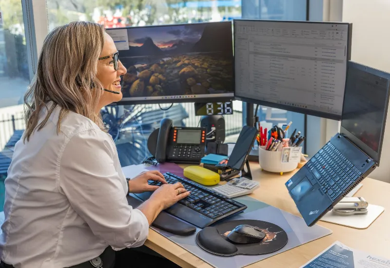 A woman at an office desk wears a headset and types on a keyboard, surrounded by dual monitors, a laptop, and office supplies.