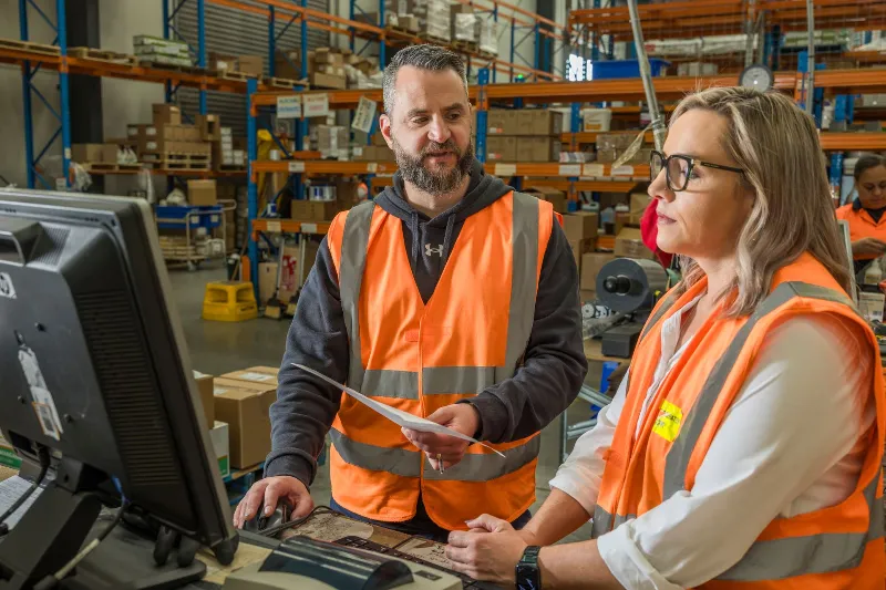 Two warehouse staff wearing orange high-visibility vests stand at a workstation, reviewing paperwork and information on a computer screen inside a busy warehouse with shelving and boxed inventory in the background.